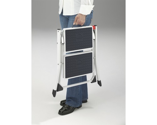 Zusammengeklappte Trittleiter mit zwei Stufen wird von einer Person getragen.