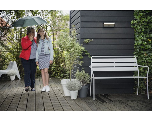 Scène de jardin avec deux personnes sous un parapluie, un banc de jardin, des plantes en pot et une applique murale sur un mur en bois.