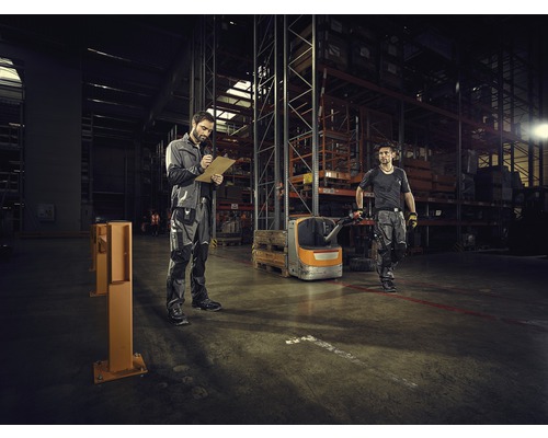 Deux travailleurs dans un entrepôt avec des étagères et un chariot élévateur. L''un prend des notes.