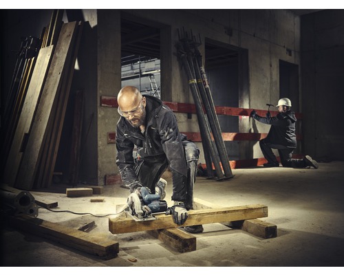 Un ouvrier du bâtiment scie une planche de bois avec une scie circulaire sur un chantier. Un autre ouvrier du bâtiment est visible en arrière-plan.