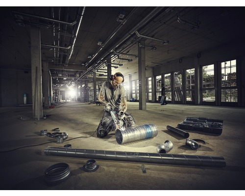 Un homme agenouillé dans une construction brute en train de travailler un tuyau avec une meuleuse d''angle, entouré d''autres tuyaux et outils.