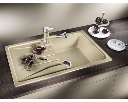 Évier de cuisine beige avec égouttoir, robinetterie, bonde à panier et aliments sur un plan de travail foncé