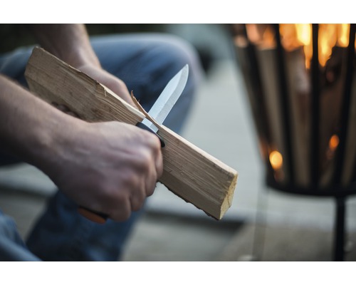 Une personne coupe du bois avec un couteau devant une vasque à feu.