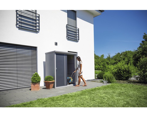 Frau betritt Gartenhaus mit Schlauchtrommel vor weißem Haus