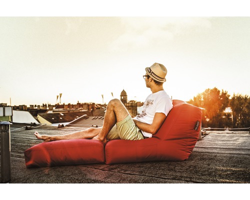 Mann mit Hut entspannt auf rotem Sitzsack mit Fußteil auf einem Dach