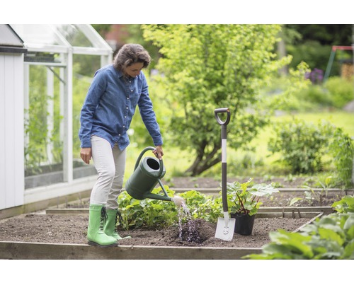 Frau giesst Pflanzen im Garten mit Giesskanne neben Spaten und Gewächshaus