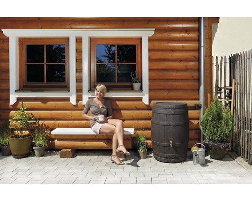 Außenbereich mit Holzwand, Fenstern, Bank, sitzender Frau, Regentonne und Pflanzen