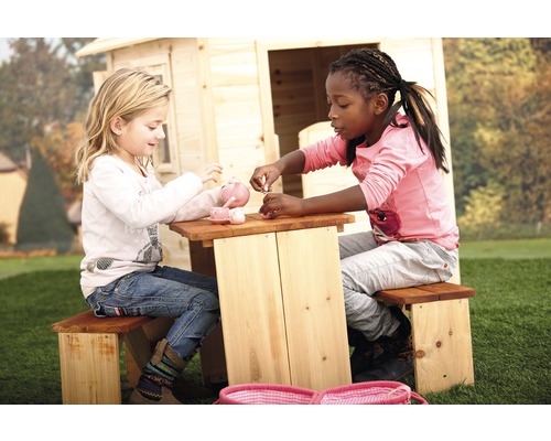 Zwei Kinder sitzen an einem Picknicktisch aus Holz und spielen mit einem Teeservice vor einem Spielhaus.