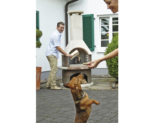Un homme met du bois dans un barbecue de jardin pendant qu''un chien regarde.