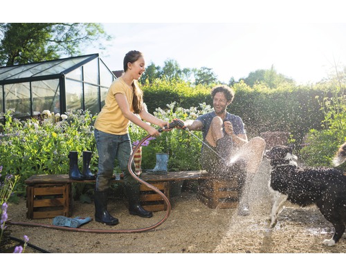 Eine junge Frau hält einen Gartenschlauch und spritzt einen Mann und einen Hund im Garten nass. Im Hintergrund befindet sich ein Gewächshaus.