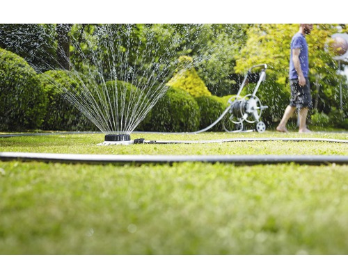 Rasensprenger bewässert einen Rasen mit einem Schlauchwagen und einer Person im Hintergrund.