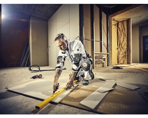 Un artisan est agenouillé dans un chantier et coupe une plaque de plâtre le long d''un niveau à bulle avec un cutter. Des outils et des câbles sont éparpillés autour.