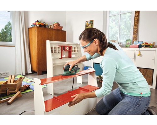 Frau schleift in einem Raum ein Regal mit einem Schleifgerät.