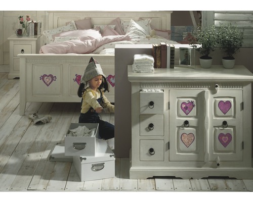 Une fille joue dans une chambre d''enfant avec un lit, une table de chevet et une commode de style campagnard.