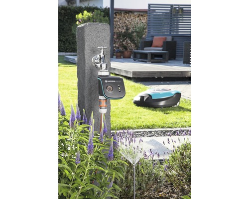 Szene mit einem Wasserhahn, einem Gardena Bewässerungscomputer, einem Rasensprenger und einem Mähroboter im Garten.