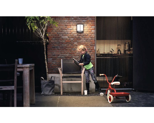 Aussenbereich mit Backsteinmauer, beleuchteter Wandleuchte, Holztisch, Stuhl und spielendem Kind