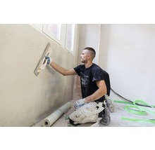 Un homme lisse un mur avec une spatule dans une pièce lumineuse.