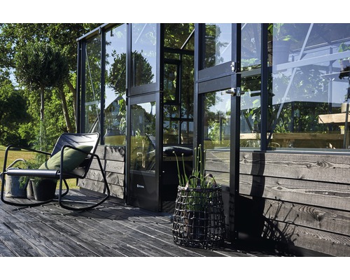 Vue extérieure d''une serre avec des murs en verre, un coin salon et des plantes décoratives sur une terrasse en bois