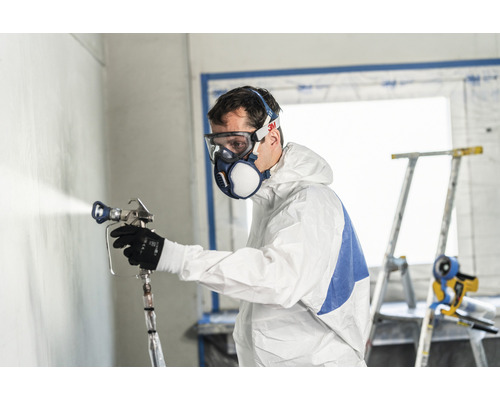 Mann mit Schutzanzug und Atemschutzmaske beim Sprühen von Farbe auf eine Wand