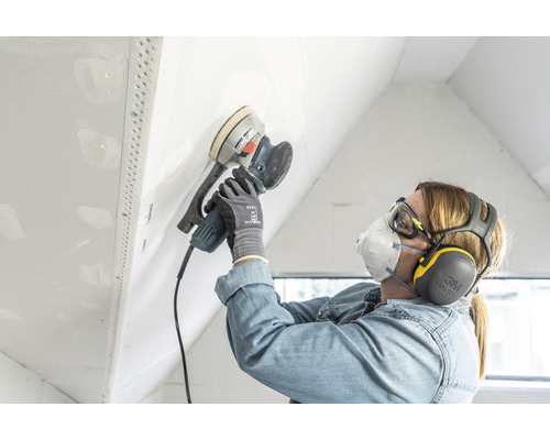 Eine Person schleift eine Wand mit einem Exzenterschleifer, während sie Schutzbrille, Atemschutzmaske und Gehörschutz trägt.