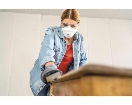 Frau schleift mit Schleifpapier ein Möbelstück, dabei trägt sie eine Schutzmaske und eine Schutzbrille
