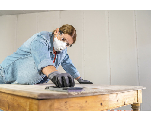 Frau schleift Tisch mit Schleifpapier, Schutzbrille und Atemschutzmaske
