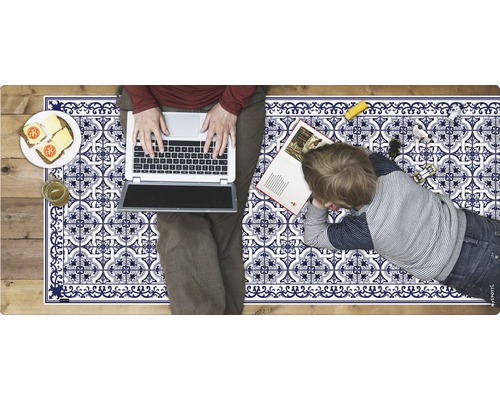 Szene mit einer Kindermatratze mit blau-weißem Fliesenmuster, einer Person mit Laptop, einem Kind mit Buch, Speisen und Spielzeug.