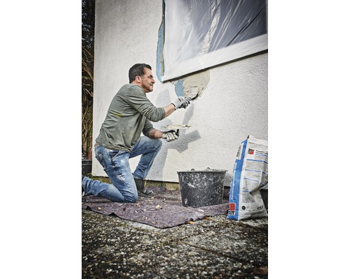 Ein Mann trägt mit einer Kelle Putz auf eine Hauswand auf. Neben ihm steht ein Eimer und ein Sack Putz.