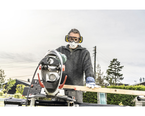 Un homme portant des protections auditives, des lunettes de protection et un masque respiratoire travaille sur une planche de bois avec une scie à onglet