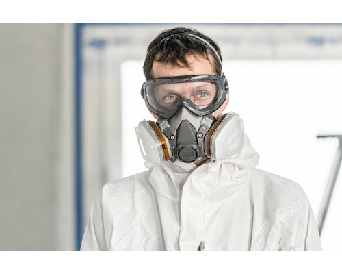 Eine Person trägt eine Schutzbrille, einen Atemschutz und einen Schutzanzug.