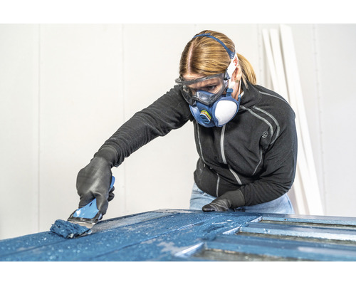 Frau mit Schutzbrille und Atemschutzmaske streicht Tür mit Pinsel