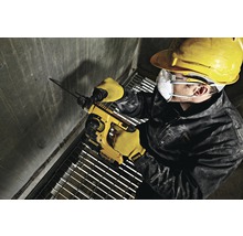 Ein Arbeiter bohrt mit einem Bohrhammer ein Loch in eine Betonwand. Er trägt Schutzkleidung wie Schutzbrille, Schutzhelm und Handschuhe.