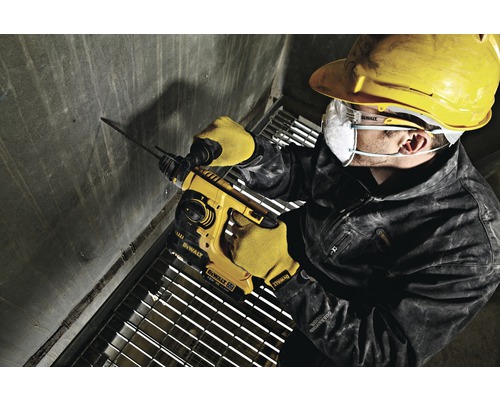 Ein Arbeiter bohrt mit einem Bohrhammer ein Loch in eine Betonwand. Er trägt Schutzkleidung wie Schutzbrille, Schutzhelm und Handschuhe.