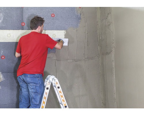 Un homme fixe des panneaux isolants à un mur avec de la colle et des outils.