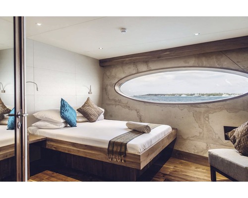 Chambre lumineuse avec lit, coussins et fenêtre ovale avec vue sur la mer