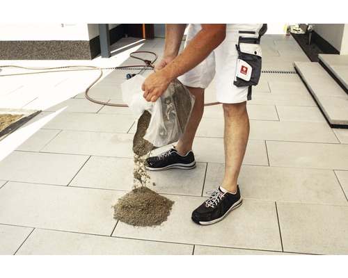 Un homme verse du sable de jointoiement sur une zone pavée à l''extérieur.