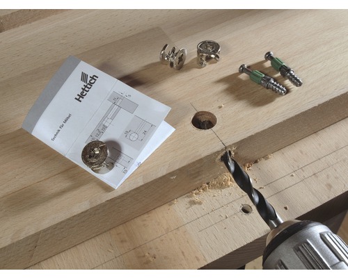 Perceuse perçant un trou dans une planche de bois à côté de connecteurs de meubles et du logo Hettich