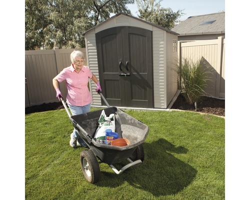 Une femme pousse une brouette contenant des outils et du matériel de jardinage devant une remise de jardin.