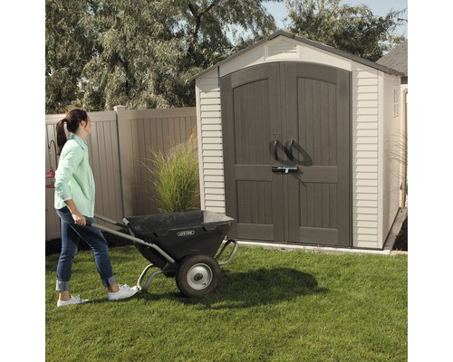Une femme pousse une brouette devant un abri de jardin.