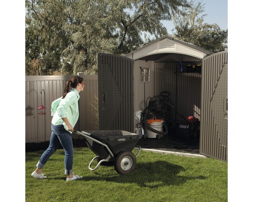Une femme pousse une brouette dans un abri de jardin avec des portes ouvertes contenant du matériel de jardinage.