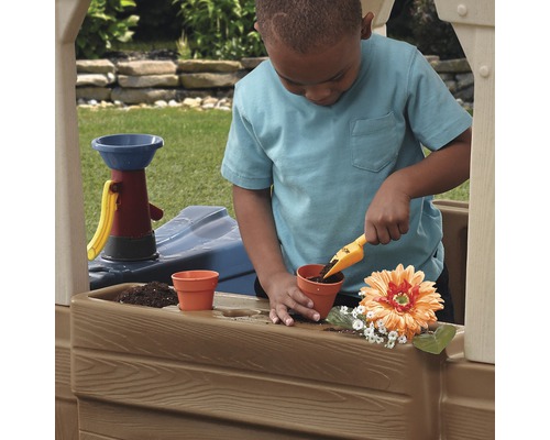 Ein Kind spielt in einem Outdoor-Spielhaus mit einer Schaufel und Blumentöpfen.