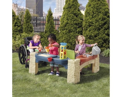 Drei Kinder spielen mit einem Spieltisch im Garten. Ein Kind sitzt im Rollstuhl.