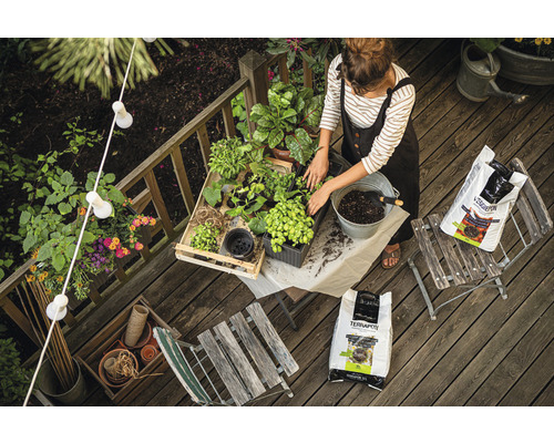 Frau bepflanzt Pflanzen auf einer Holzterrasse mit Pflanzerde, umgeben von Gartengeräten und Dekoration