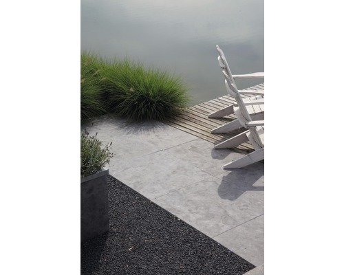 Szene mit Gartengestaltung, inklusive Terrasse mit grauen Steinplatten, Kiesfläche, Bepflanzung und weißen Gartenstühlen am Wasser