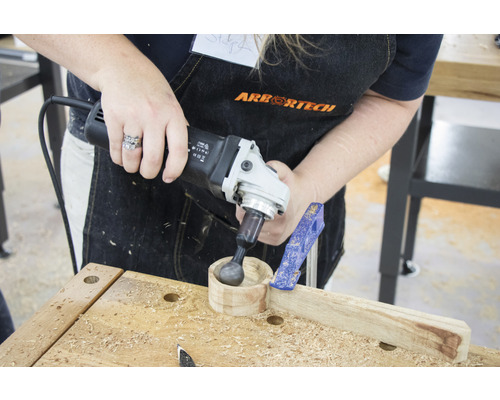 Eine Person bearbeitet mit einem Elektrowerkzeug eine kleine Holzschale auf einer Werkbank. Eine Zwinge fixiert das Werkstück.