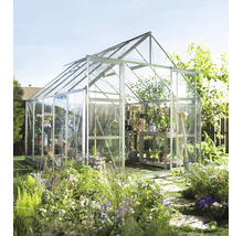 Serre autonome en aluminium avec parois vitrées et lucarne dans un jardin ensoleillé avec parterres de fleurs et outils de jardinage.