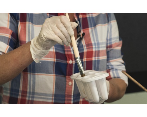 Une personne gantée trempe un pinceau dans un récipient de peinture.