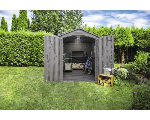 Jardin avec remise à outils ouverte contenant des outils de jardinage, un vélo et du bois.