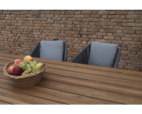 Scène de mobilier de jardin avec table en bois, deux chaises avec coussins et une corbeille de fruits devant un mur de briques