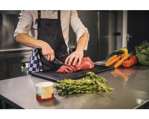 Une personne coupe de la viande sur une planche à découper avec des légumes en arrière-plan.